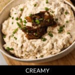 A bowl of creamy caramelized onion dip, perfect for any occasion, is topped with chopped chives and more caramelized onions, all elegantly placed on a wooden board beside slices of crusty bread.