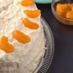 A frosted Pig Cake garnished with mandarin orange slices sits on a glass platter, alongside a bowl of mandarin segments and a plate with a slice of the cake.