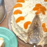 A round Pig Cake with yellow frosting, topped with mandarin orange slices, is partially sliced and placed on a clear glass plate with a green plate next to it.