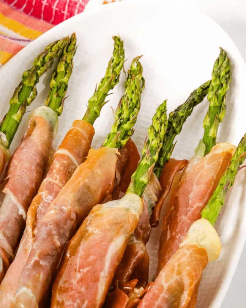 A plate of succulent, prosciutto-wrapped asparagus spears, neatly arranged on a white surface with a vibrant cloth in the background.