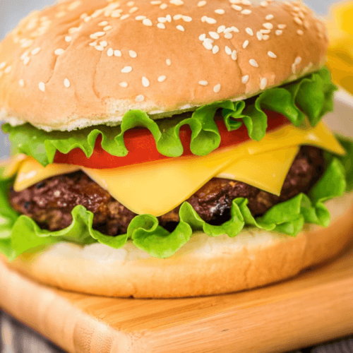A baked hamburger with lettuce, tomato, and cheese in a sesame seed bun, placed on a wooden board.