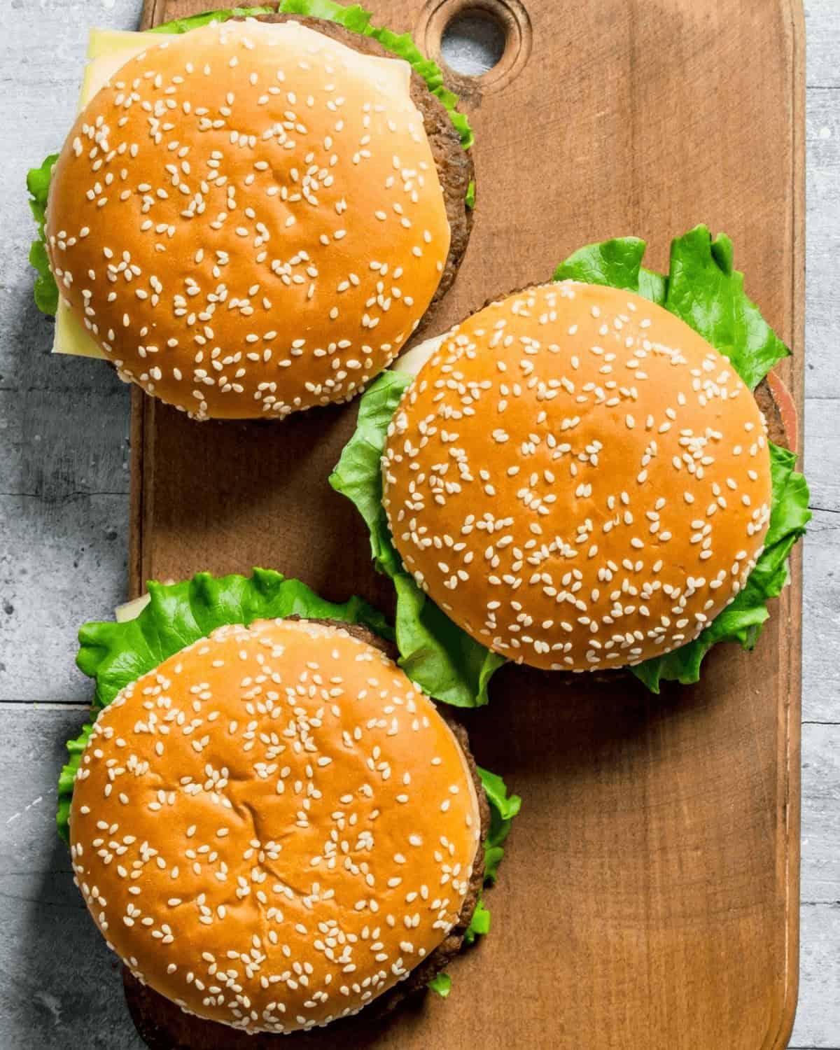 Three perfectly baked hamburgers with sesame seeds and fresh lettuce sit enticingly on a wooden board.