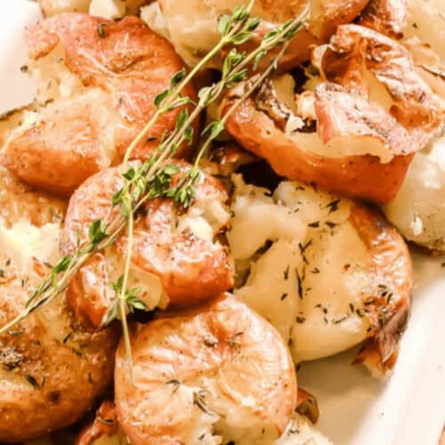 Close-up of roasted smashed potatoes garnished with fresh thyme on a white plate. The potatoes have a golden-brown, crispy texture.