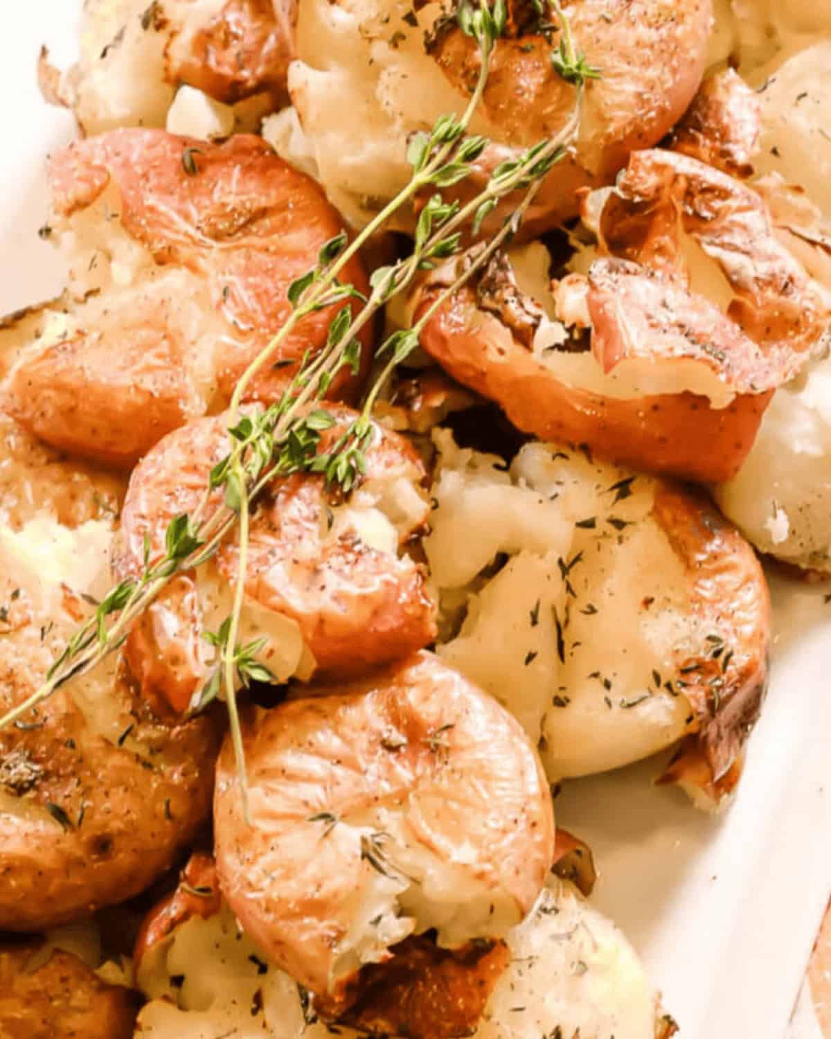 Close-up of roasted smashed potatoes garnished with fresh thyme on a white plate. The potatoes have a golden-brown, crispy texture.