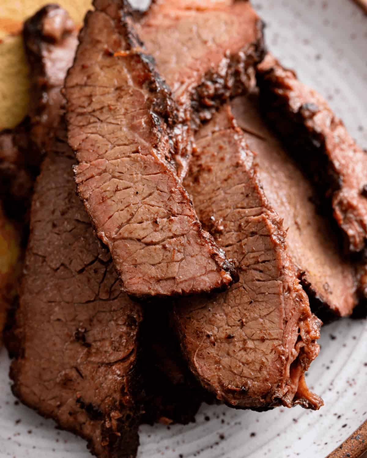 Close-up of several slices of grilled, smoked brisket, seasoned and well-cooked, on a white plate.