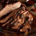 Smoked brisket sliced on a wooden cutting board, with a small fork and a dish of barbecue sauce in the background.