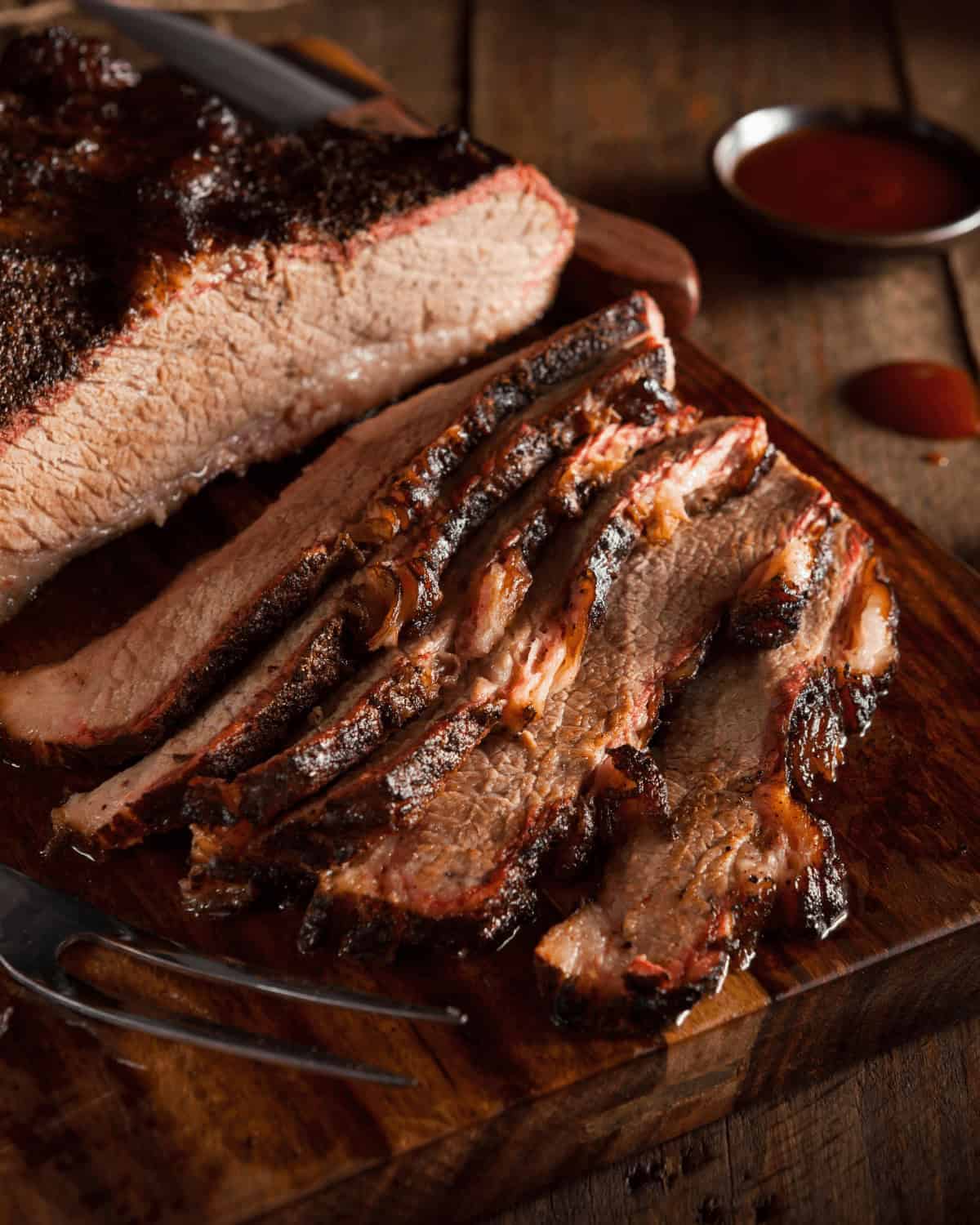 Smoked brisket sliced on a wooden cutting board, with a small fork and a dish of barbecue sauce in the background.