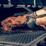 Person wearing black gloves carefully placing a large piece of smoked brisket on a grill rack.