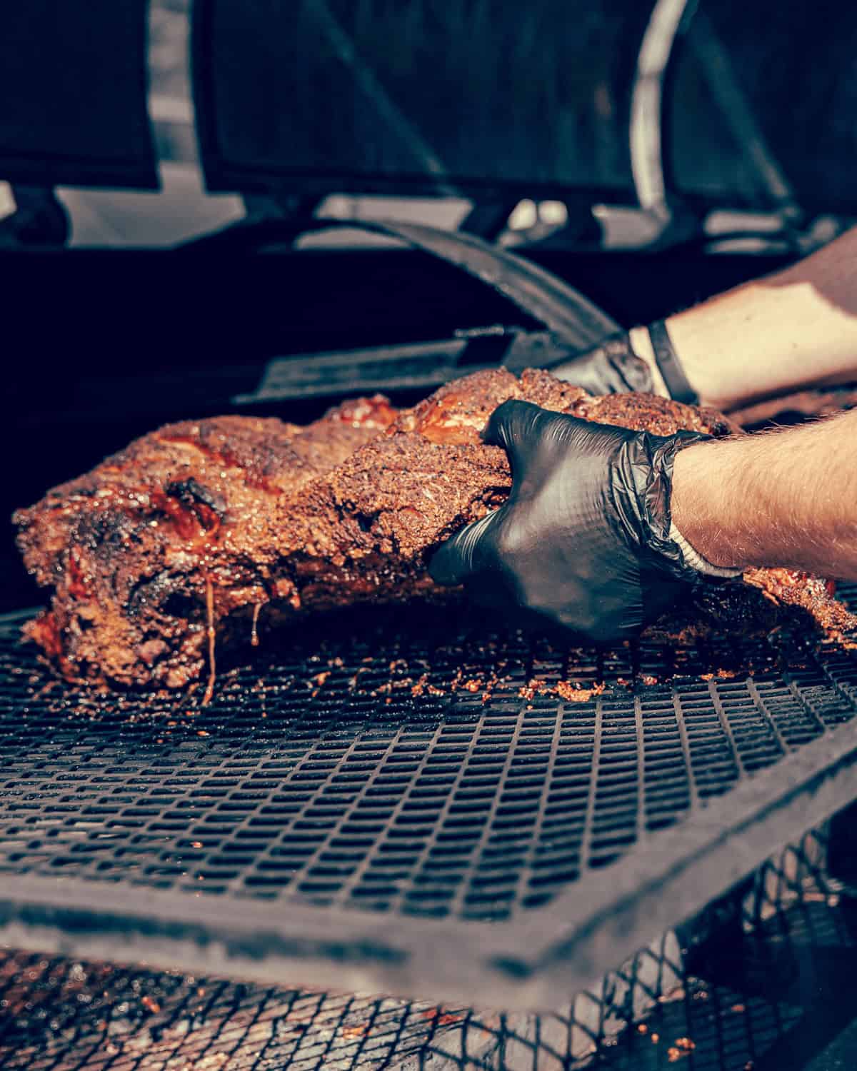 Person wearing black gloves carefully placing a large piece of smoked brisket on a grill rack.