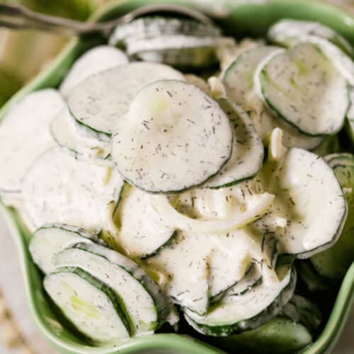 A bowl of creamy cucumber onion salad with fresh dill, served in a light green bowl with a silver spoon.