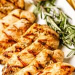 Lemon rosemary chicken breast pieces with grill marks, garnished with sprigs of rosemary and a charred lemon, served on a white rectangular plate.