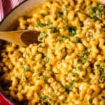 A red pot filled with veal cheeseburger macaroni and cheese topped with chopped herbs, with a wooden spoon resting inside.