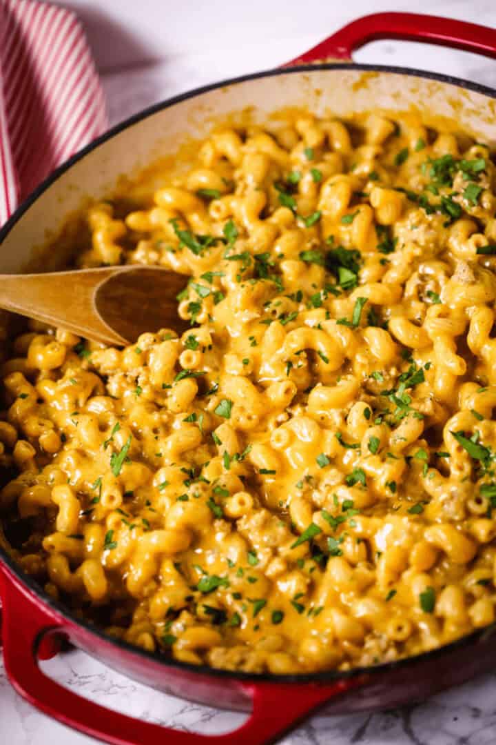A red pot filled with veal cheeseburger macaroni and cheese topped with chopped herbs, with a wooden spoon resting inside.