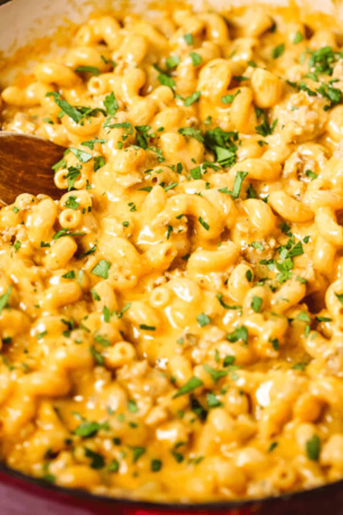A close-up of creamy cheeseburger macaroni sprinkled with fresh herbs, with a wooden spoon partially visible on the left side of the dish.