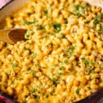 A red pan brimming with creamy cheeseburger macaroni, the dish is garnished with chopped herbs and accompanied by a wooden spoon resting inside.