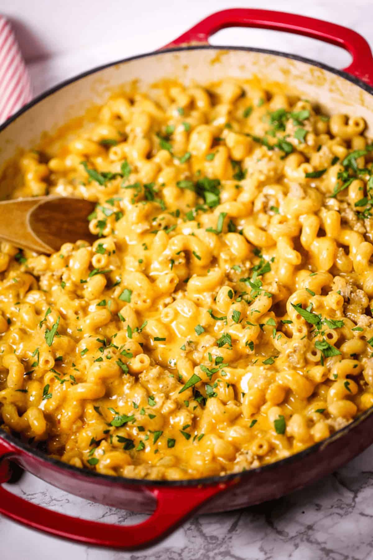 A red pan brimming with creamy cheeseburger macaroni, the dish is garnished with chopped herbs and accompanied by a wooden spoon resting inside.