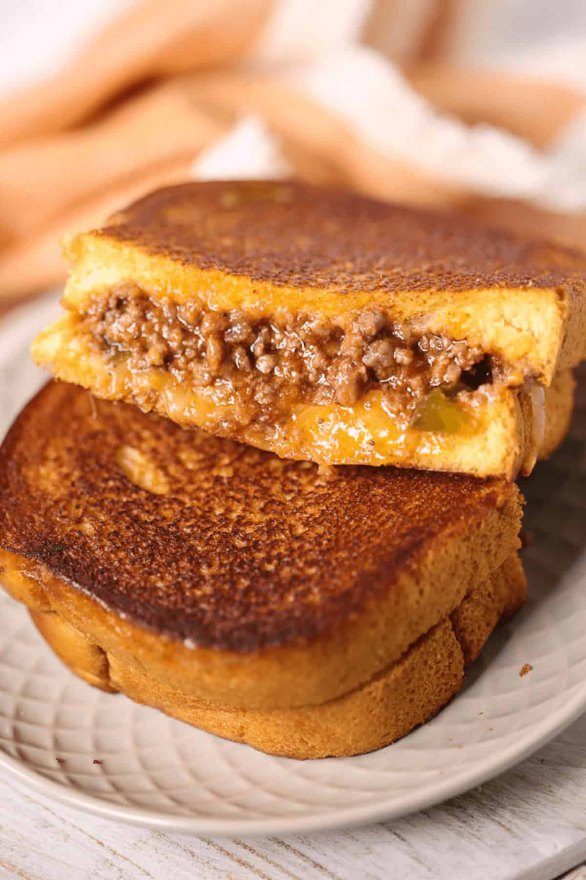 A Sloppy Joe Grilled Cheese sandwich, brimming with melted cheese and savory ground beef filling, is expertly grilled, cut in half, and elegantly stacked on a plate.