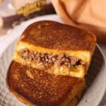 A Sloppy Joe grilled cheese sandwich, with a filling of seasoned ground beef and gooey melted cheese, is partially sliced and artfully stacked on a plate. In the background, a bread package adds the finishing touch to this deliciously messy creation.