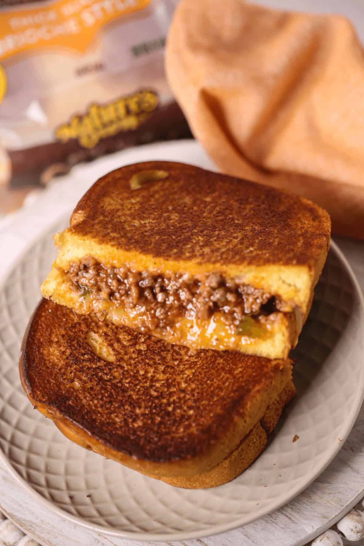 A Sloppy Joe grilled cheese sandwich, with a filling of seasoned ground beef and gooey melted cheese, is partially sliced and artfully stacked on a plate. In the background, a bread package adds the finishing touch to this deliciously messy creation.