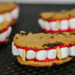 Vampire cookie creations featuring mouths with red icing, marshmallow teeth, and almond slivers as fangs, all artfully arranged on a black surface.