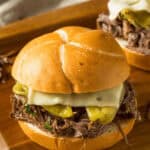 A Mississippi Pot Roast sandwich with shredded beef, melted cheese, and pickles on a bun, placed on a wooden board.