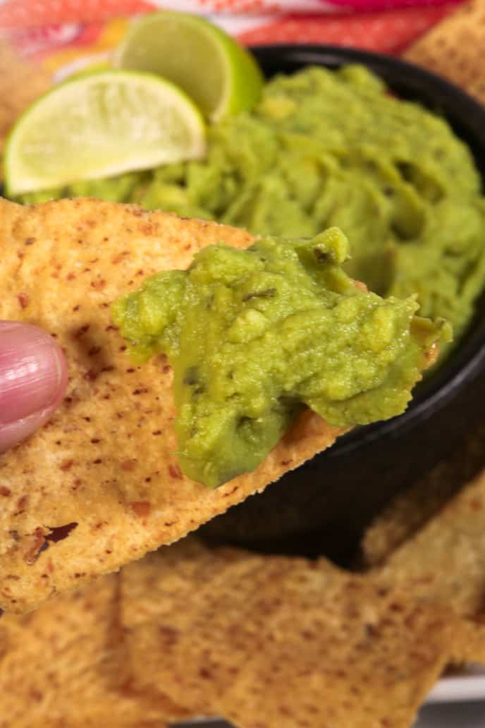 Guacamole on a tortilla chip, with black bowl of guacamole and limes in the background. 
