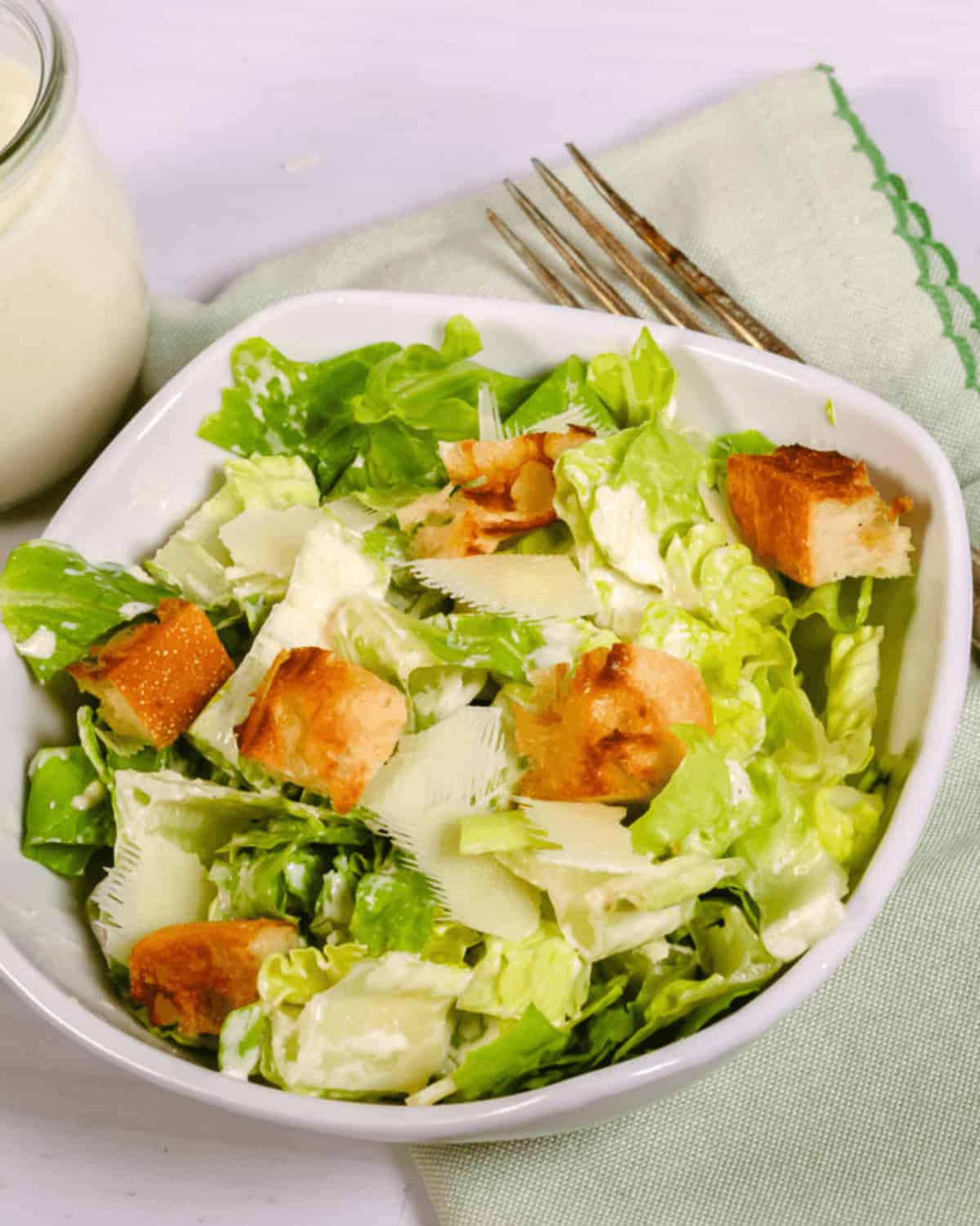 Bowl of the best Caesar salad with croutons and shaved parmesan cheese, accompanied by a fork and dressing on the side.