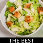 Best Caesar salad with croutons and shaved cheese in a white bowl, with dressing on the side.