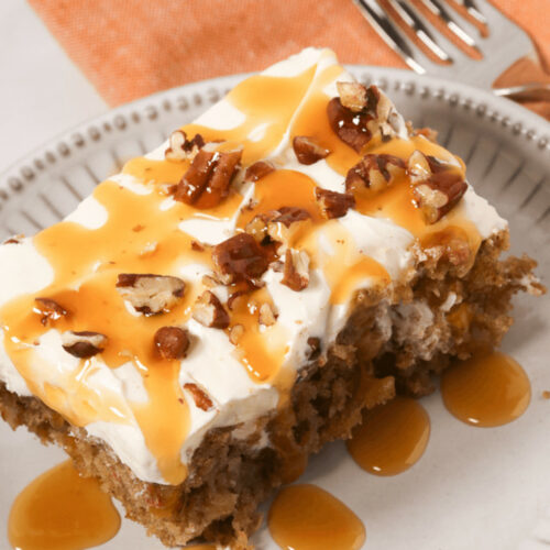 A piece of carrot poke cake topped with cream cheese frosting, caramel drizzle, and chopped pecans, served on a decorative plate.
