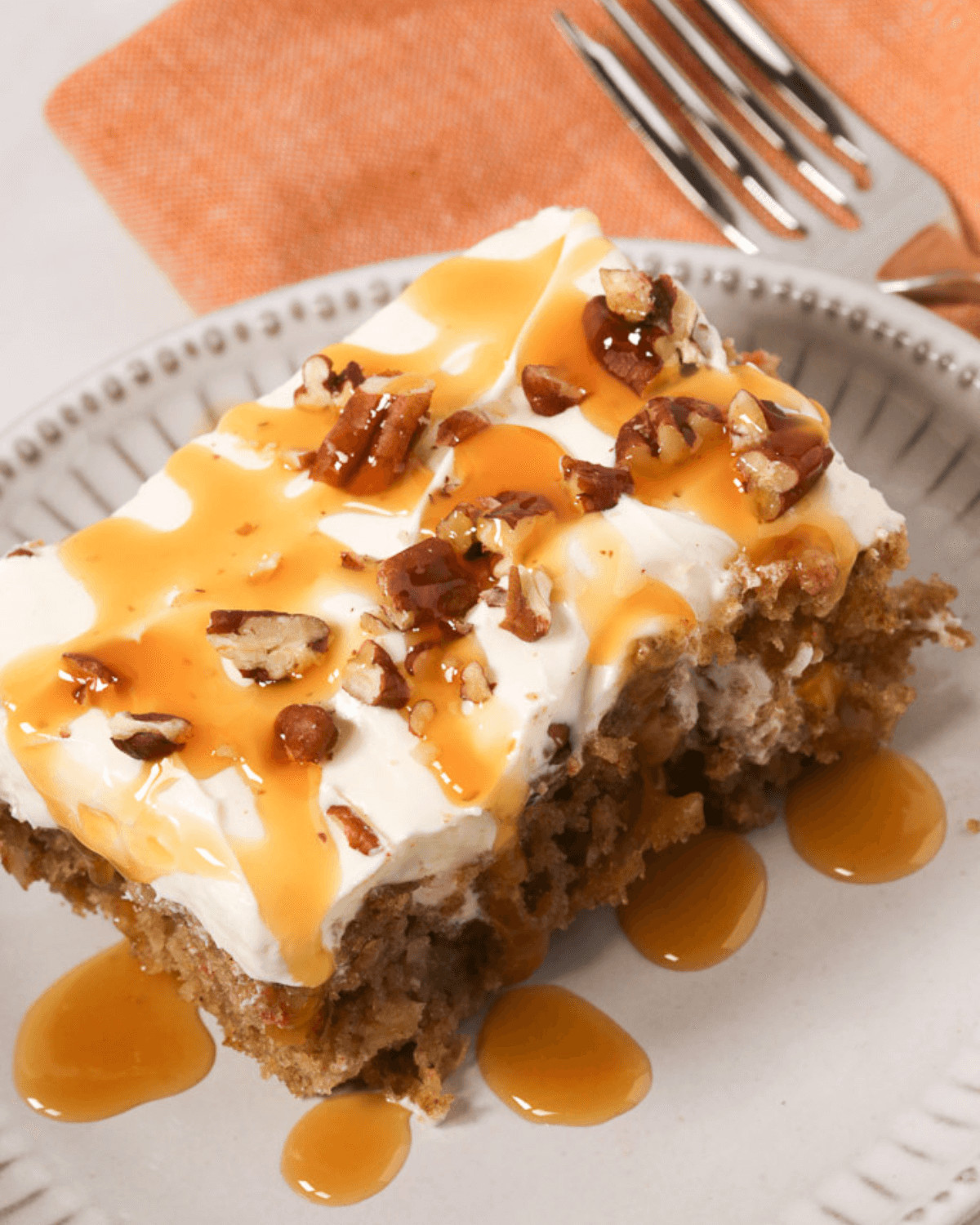 A piece of carrot poke cake topped with cream cheese frosting, caramel drizzle, and chopped pecans, served on a decorative plate.