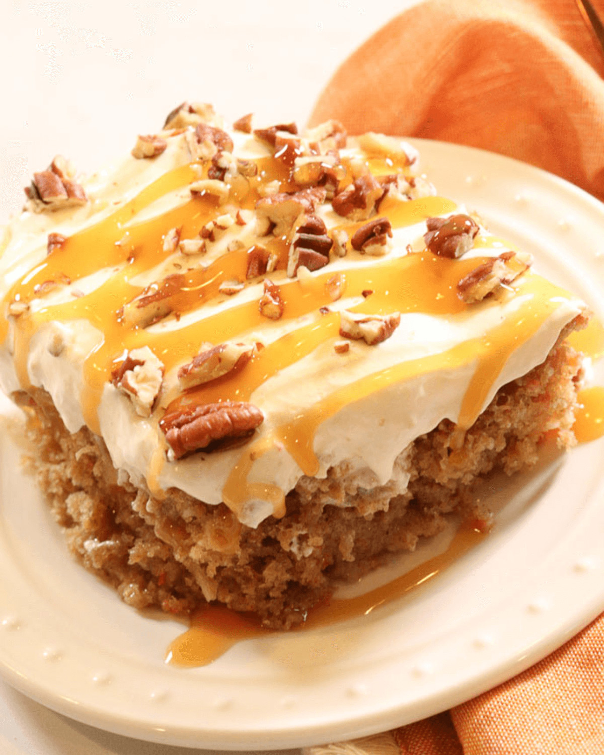 Slice of carrot poke cake with cream cheese frosting, caramel drizzle, and chopped pecans on a white plate.