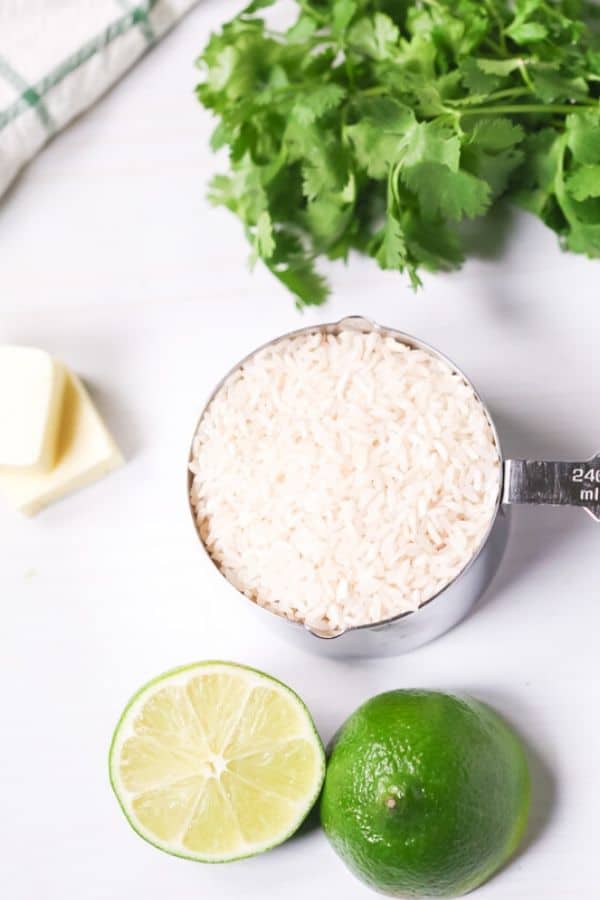Uncooked rice in a measuring cup with cilantro, limes and butter on the side