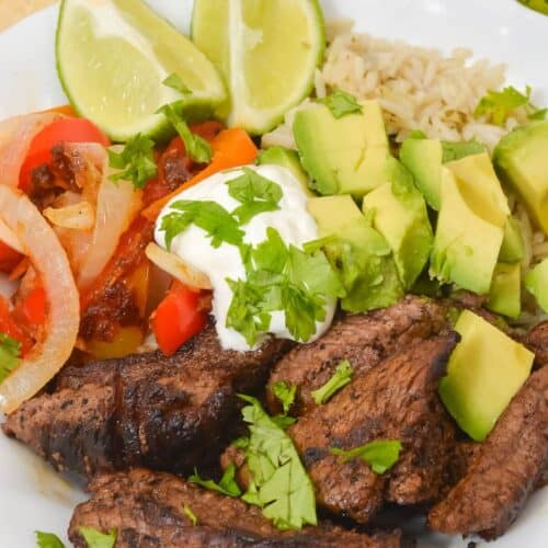 A Steak and Rice Bowl featuring grilled steak, sliced avocado, brown rice, sautéed peppers and onions, topped with sour cream and cilantro, served with lime wedges and tortilla chips in the background.