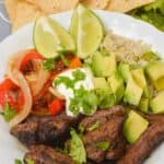 A Bowl featuring meat, avocado cubes, rice, sautéed onions and peppers, lime wedges, sour cream, and fresh cilantro, served with tortilla chips in the background.