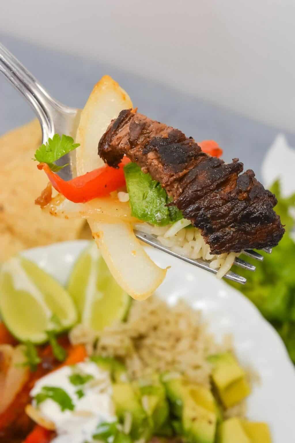 A fork holding grilled steak, onion, red pepper, and green pepper above a vibrant dish with avocado, sour cream, and lime wedges.