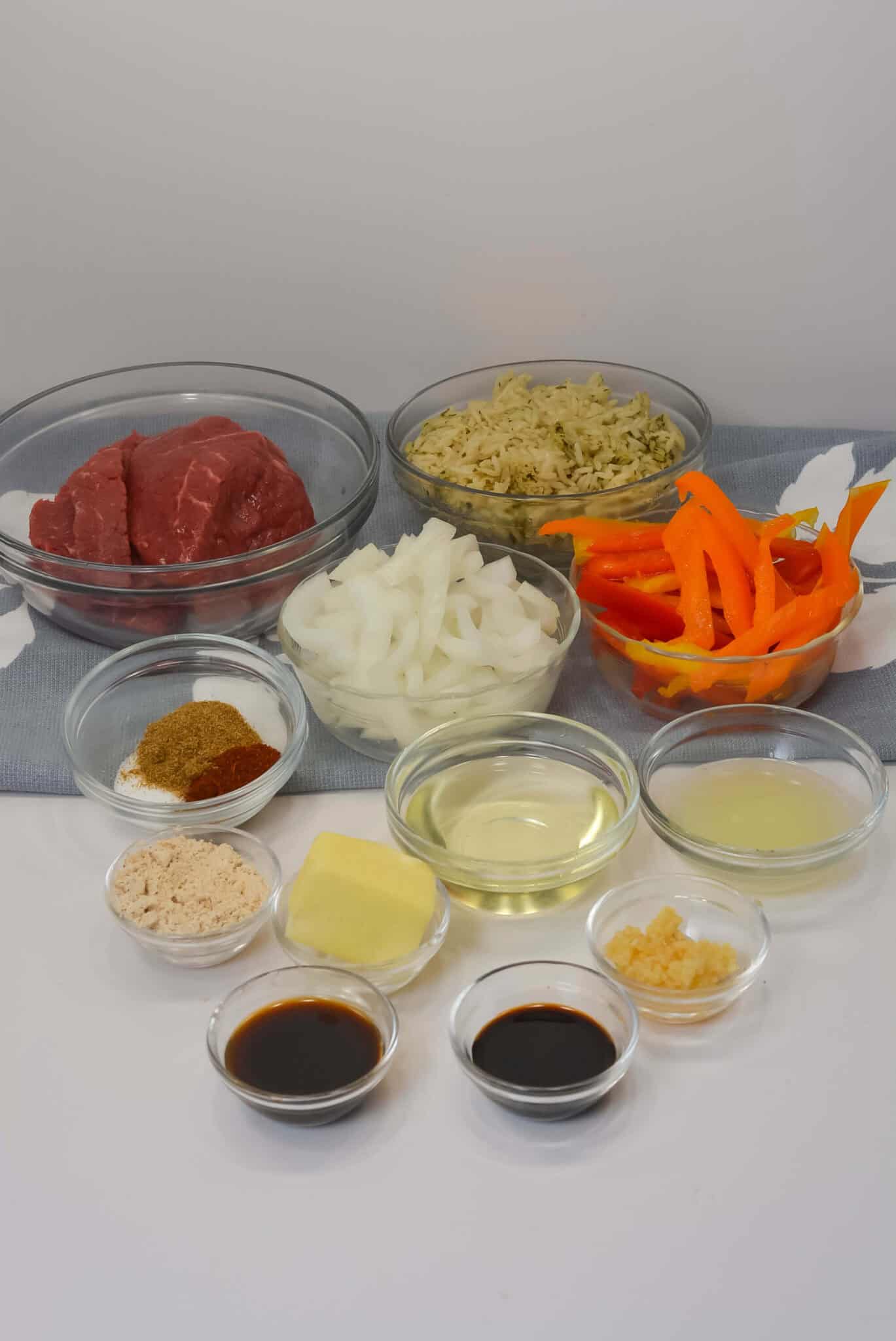 Various ingredients  are displayed in small bowls, including raw beef, sliced onions, orange bell peppers, cooked rice, liquid seasonings, spices, minced garlic, a pat of butter, and a light-colored liquid.