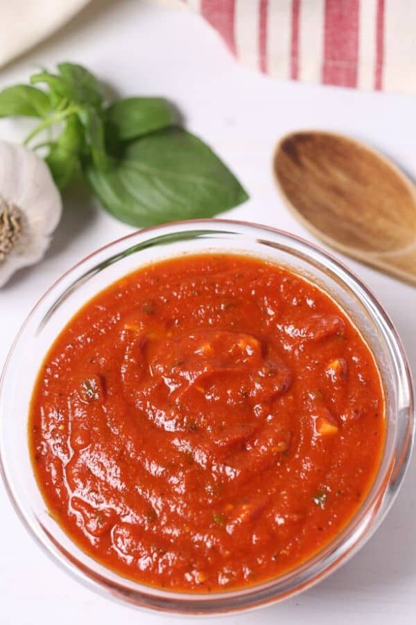 Homemade pizza sauce in a glass bowl with garlic and basil on the side