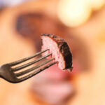 A piece of grilled, medium-rare steak on a fork with a blurred background.