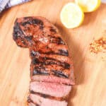 Grilled London broil sliced and served on a wooden board with lemon halves and spices.