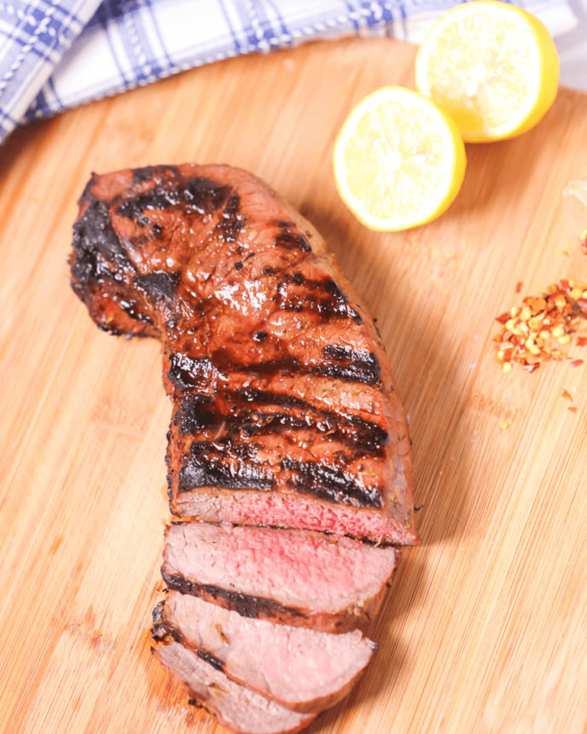 Grilled London broil sliced and served on a wooden board with lemon halves and spices.