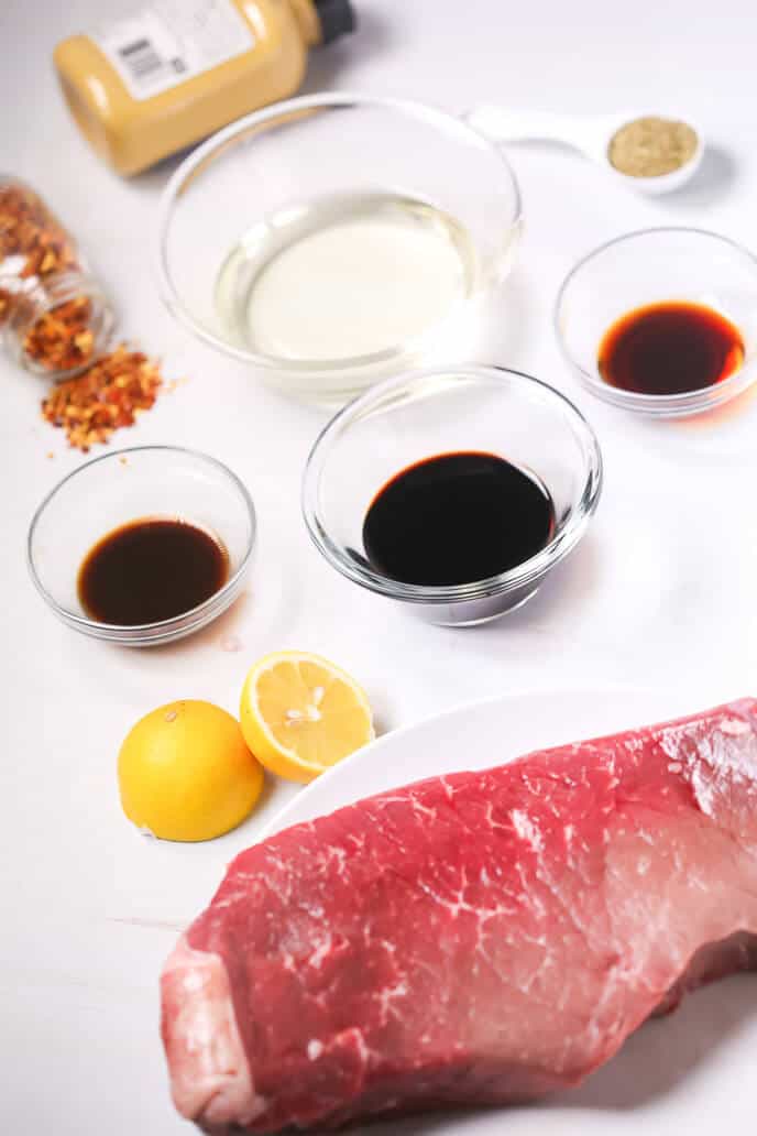 Various ingredients for cooking, including a grilled London broil, lemon, and several condiments in bowls, arranged on a kitchen surface.