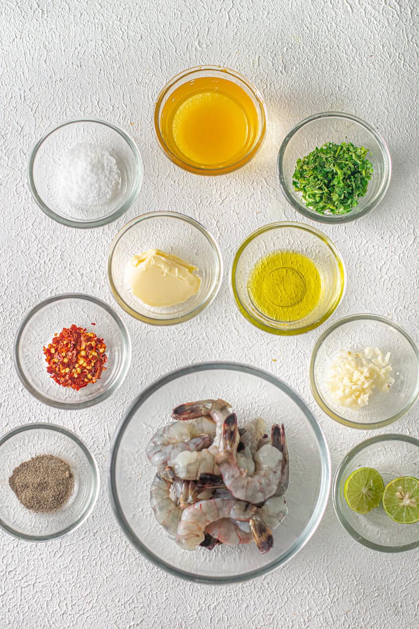 Various ingredients for a Shrimp Scampi without Wine recipe neatly arranged in separate bowls on a textured white surface.