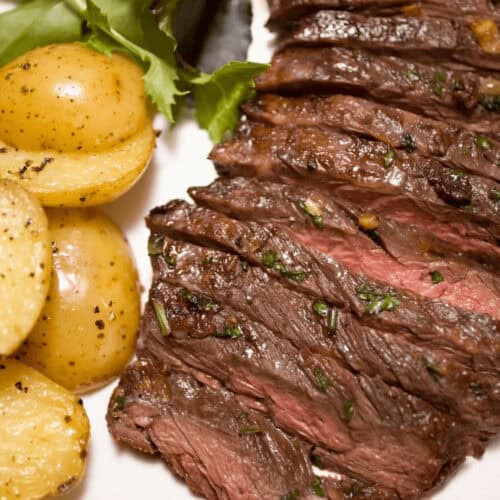Sliced grilled skirt steak with herbs, served alongside boiled potatoes and a green salad, presented on a white plate.