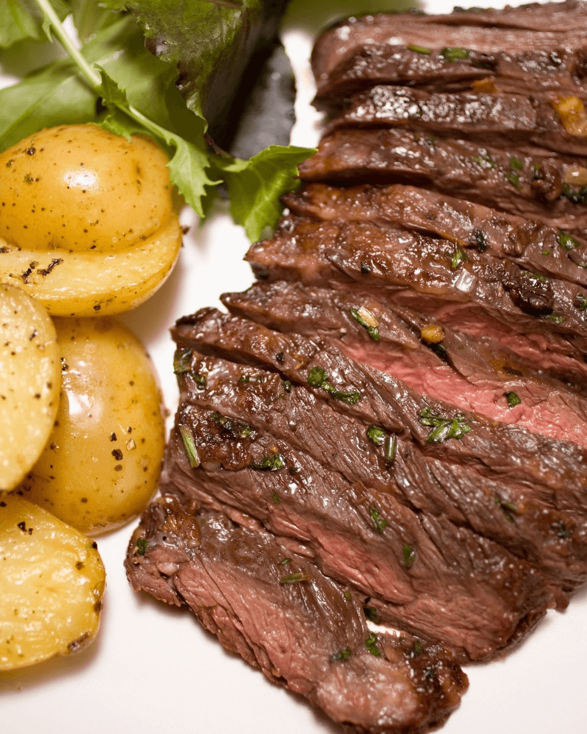 Sliced grilled skirt steak with herbs, served alongside boiled potatoes and a green salad, presented on a white plate.