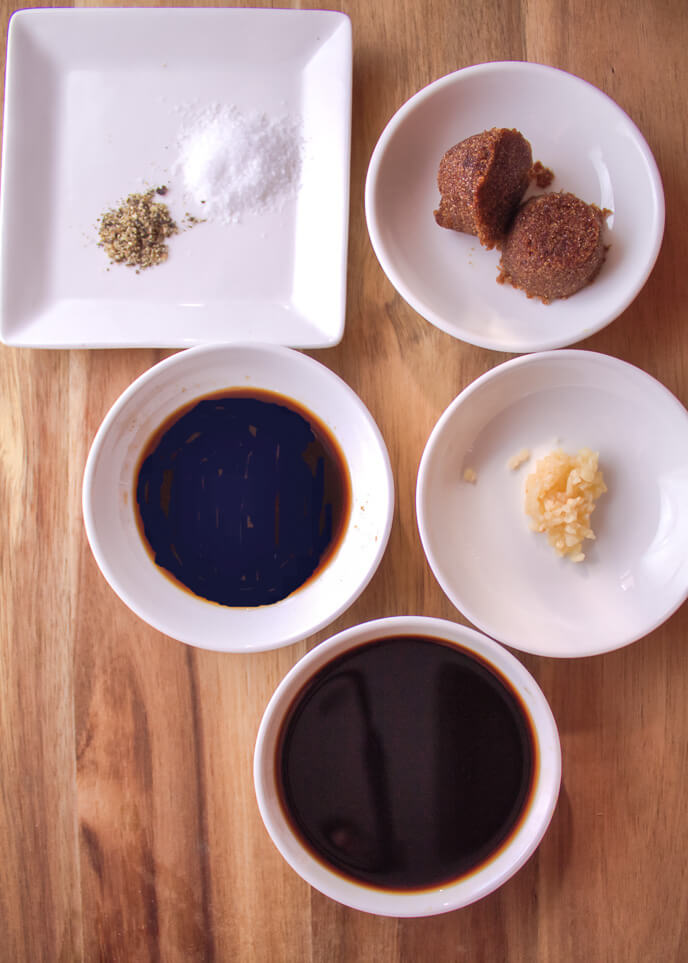Overhead view of various condiments on a wooden table, accompanying grilled skirt steak, including bowls of salt, pepper, soy sauce, brown sauce, and minced garlic.
