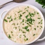 A bowl of creamy Instant Pot risotto garnished with chopped parsley, placed on a light surface. A spoon and some parsley leaves are in the background.