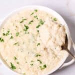 A bowl of creamy side dish topped with chopped parsley, with a spoon elegantly lifting a portion from the side.