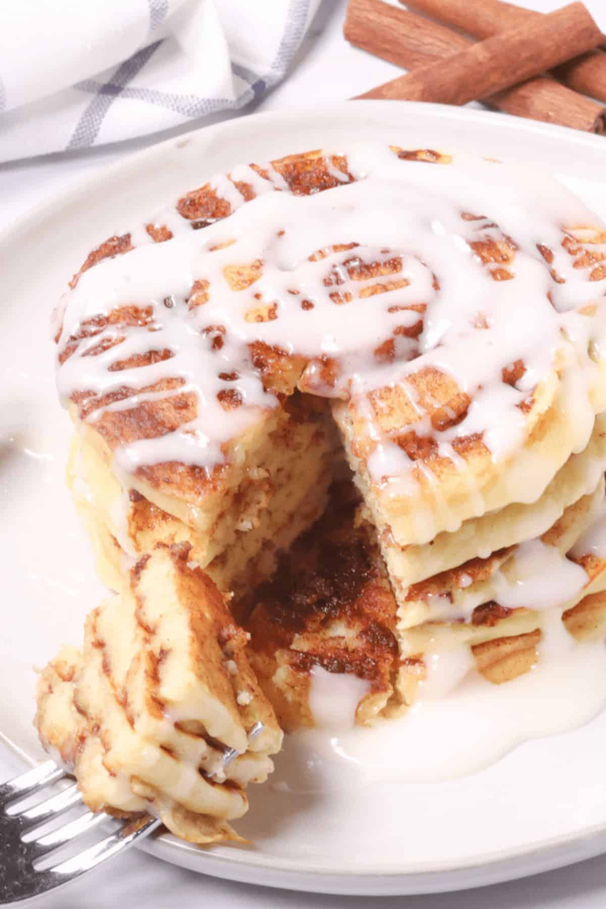 A stack of cinnamon swirl pancakes, with an icing drizzle and a portion artfully cut out on a fork, rests invitingly on a white plate.