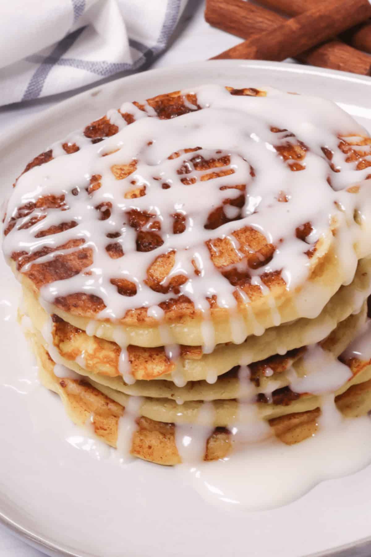 A stack of four cinnamon swirl pancakes topped with white icing sits invitingly on a plate, with a striped cloth and cinnamon sticks nestled in the background.