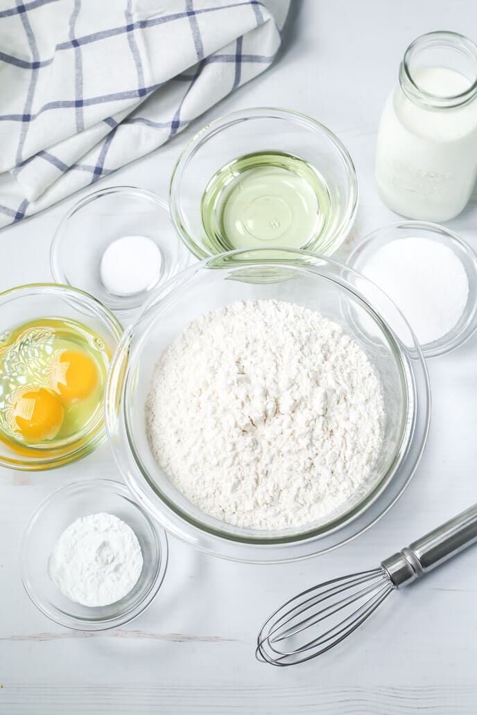 Baking ingredients on a table include flour, eggs, milk, oil, sugar, baking powder, and salt—all set for crafting delicious cinnamon swirl pancakes. A whisk and a striped cloth are nearby.